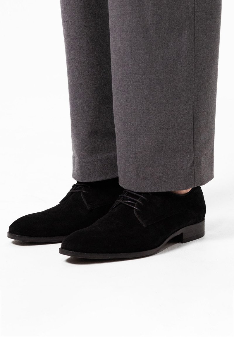 Zwarte suede salonschoenen met een ronde neus, vetersluiting en een lage blokhak, gecombineerd met grijze maatwerkbroek.