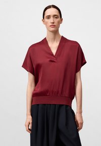 Blusa di seta bordeaux con scollatura a V e maniche corte, caratterizzata da una vita arricciata e una texture liscia. Abbinata a pantaloni neri larghi.