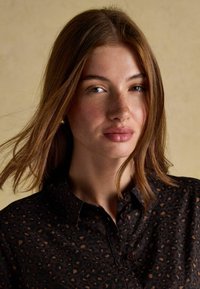 Young woman with straight brown hair, wearing a dark leopard-print button-up shirt, looking confidently at the camera against a beige background.