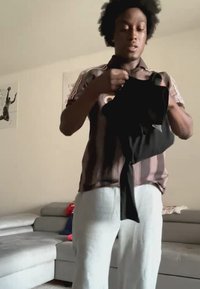 Giovane con capelli ricci si trova in un soggiorno, tiene e osserva un indumento nero, indossando una camicia a righe e pantaloni chiari.