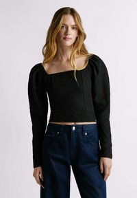 Top cropped nero con texture, maniche lunghe a sbuffo e scollatura quadrata, indossato con jeans di denim blu scuro.