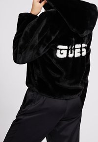 Sort hættetrøje i imiteret pels med crop-top-design, med hvid "GUESS"-tekst på ryggen. Blød tekstur og stor hætte.