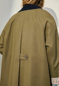 Personne portant un trench-coat vert olive avec un col sombre et des poignets à motifs, vue de dos sur un fond neutre.