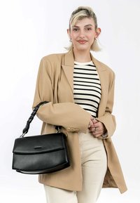 Czarna skórzana torebka z plecioną rączką, prostokątnym kształtem i klapą. Modelka w beżowej marynarce i w paskowanym topie.