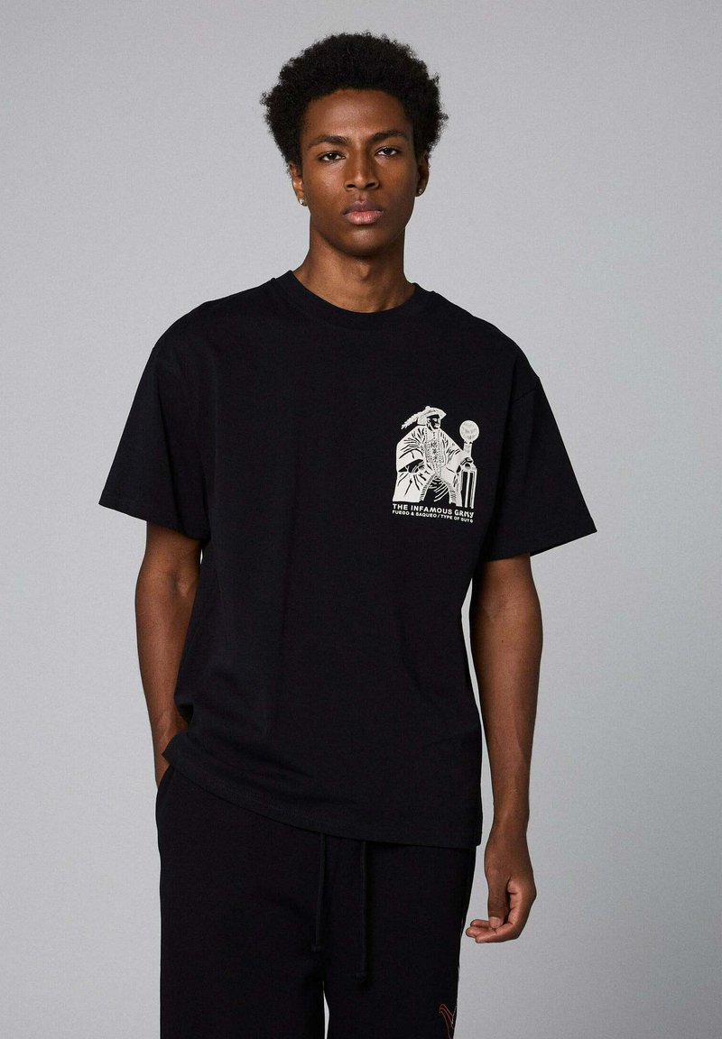 Camiseta de algodón negra con un diseño gráfico en blanco en el área del pecho izquierdo. Mangas cortas y un corte relajado, textura mínima.
