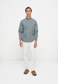 Geruite overhemd in donker groen en wit, gecombineerd met witte broek en bruine suède schoenen. Model staat tegen een effen achtergrond.