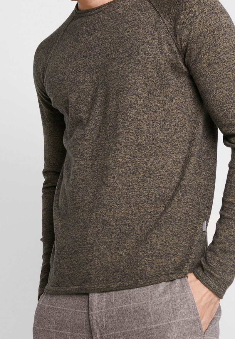 Chemise à manches longues vert foncé en tissu tricoté texturé. Dispose de manches raglan et d'un col rond. Associée à un pantalon gris clair à motifs.