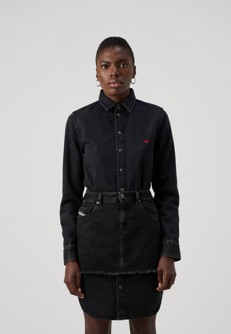Zwarte denim blouse met knoopsluiting, lange mouwen en een klein rood logo. Gecombineerd met een zwarte denim rok met een rafelige zoom.