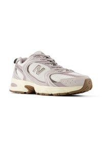 Αθλητικό παπούτσι ανοιχτού γκρι με υλικά από δίχτυ και σουέντ, διαθέτει λευκή ενδιάμεση σόλα, καφέ λαστιχένια εξωτερική σόλα και εμφανές λογότυπο "N" στο πλάι.