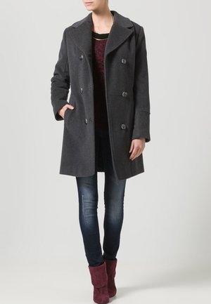 Femme portant un manteau gris foncé à double boutonnage, un jean foncé et des bottines bordeaux, debout avec une main dans la poche.