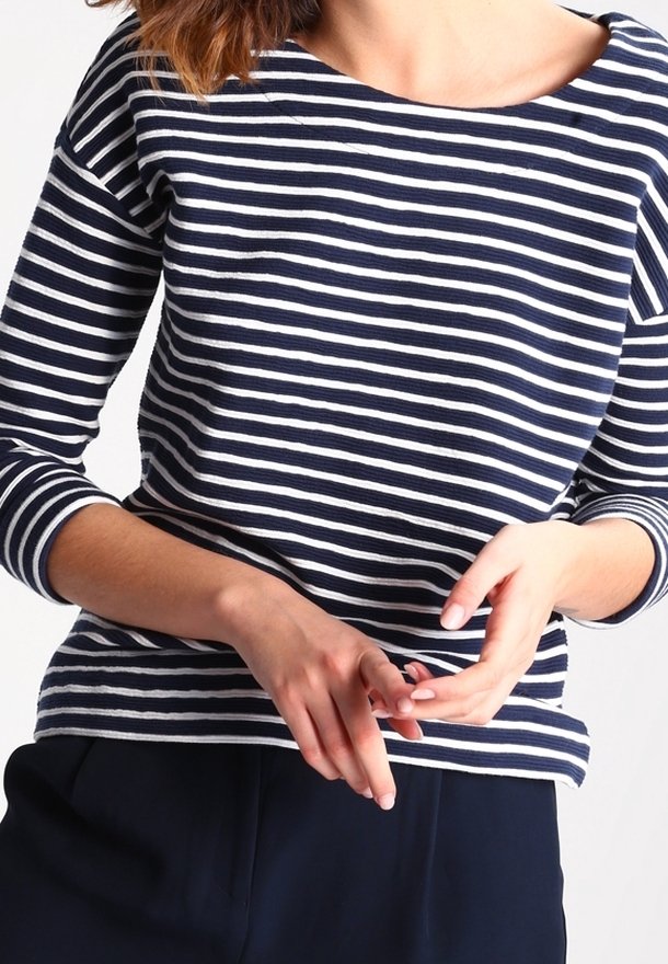 Femme portant un haut à manches longues rayé bleu marine et blanc avec un pantalon sombre, les mains doucement jointes à la taille, fond clair.