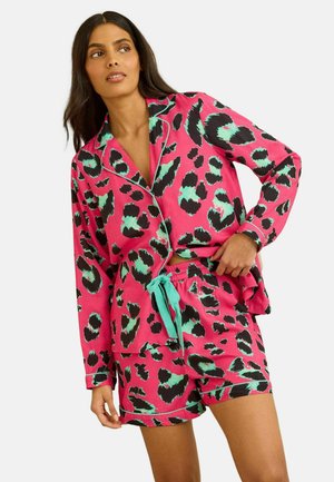 Naine kannab erkroosa ja türkiissinise leopardimustriga pikkade varrukatega särki ja sobivaid lühikesi pidžaamapükse, poseerides käega vööjoone lähedal.