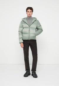 Jachetă pufoasă verde deschis, cu fermoar și glugă, asortată cu un pulover gri cu guler înalt, pantaloni negri și adidași negri. Textură netedă, design matlasat.