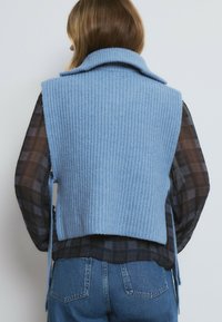 Chaleco de punto azul claro con una textura acanalada y un gran cuello, superpuesto a una camisa de cuadros oscuros, visto desde la parte posterior.