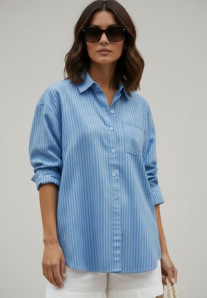 Vrouw draagt een oversized lichtblauw gestreept overhemd, zwarte zonnebril en witte short, met een witte touwen tas in haar hand.