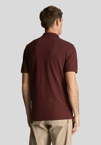 Polo color mogano con maniche corte, tessuto tinta unita, collo standard e orlo dritto, indossata con pantaloni chiari.