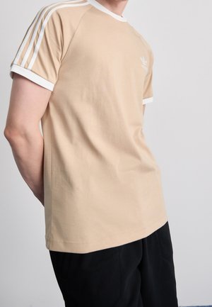 Beige bomulds t-shirt med hvide detaljer. Har tre hvide striber på hver skulder og et lille logo på brystet.