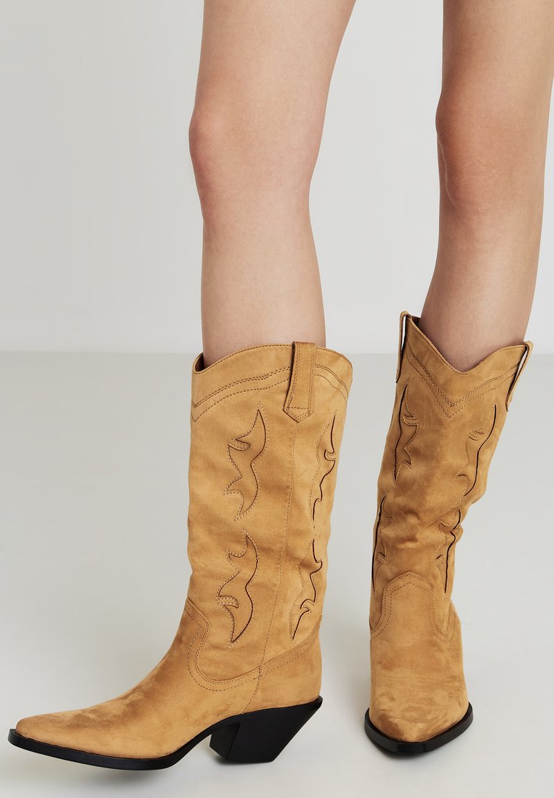 Stivali da cowboy in suede marrone con punte appuntite, motivi di cucitura intricati e tacco nero curvo. L'asta presenta ritagli decorativi.