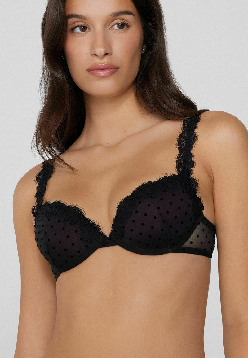 Schwarzer BH mit Spitzenrand, der aus gepunktetem Netzstoff besteht, mit tiefen Cups und verstellbaren Trägern. Das Design umfasst gewellte Kanten für zusätzliche Details.