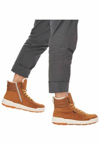 Braune Wildleder-Hochschaftstiefel mit einem weißen Futter aus Kunstfell, orangen Schnürsenkeln, einem seitlichen Reißverschluss und einer weißen Sohle, vor dunklen Hosen.
