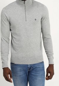 Uomo che indossa un maglione grigio chiaro con zip a quarto e un piccolo logo di pinguino sul petto, con jeans blu, in piedi su uno sfondo neutro.