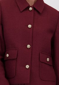 Vrouwentextuur bordeaux rode jas met goudkleurige decoratieve knopen en klepzakken, voorzien van een klassiek kraag en een gestructureerde pasvorm.
