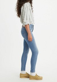 Vaqueros ajustados de mezclilla azul claro, con bolsillos traseros. Combinados con una camisa blanca a rayas y zapatos estilo mocasín de color beige claro.