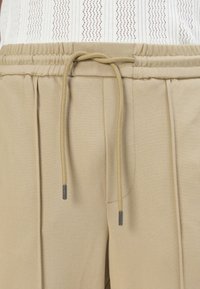 Beige Jogginghose mit glatter Textur, elastischem Bund und einem dezenten Faltenelement auf der Vorderseite.