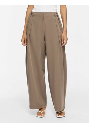 Femme portant un pantalon marron taille haute à jambes larges avec des plis devant et des sandales blanches, debout sur un fond blanc uni.