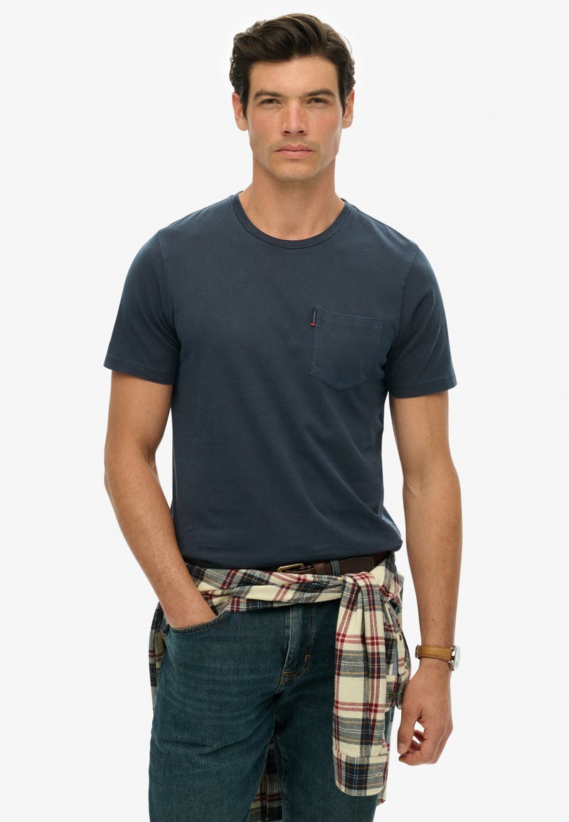 Camiseta de manga corta azul marino con un bolsillo frontal, combinada con jeans azules y una camisa de cuadro de franela atada a la cintura.