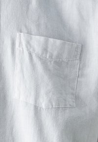 Camisa de lino blanca con un pequeño bolsillo de parche en el lado izquierdo del pecho. La tela es texturizada con un aspecto suave y ligeramente arrugado.