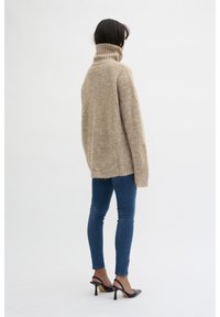 Femme de dos portant un pull oversize beige à col roulé, un jean skinny bleu avec des zips, et des chaussures à talons noires à lanières.