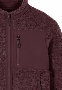 Giacca in pile bordeaux con sezioni in tessuto liscio, colletto alto e tasche con cerniera. Materiale in pile strutturato sulle maniche e sulla parte inferiore del corpo.