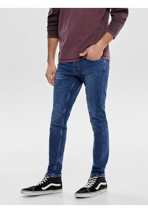 Jonge man die strakke blauwe jeans draagt, zwarte hoge sneakers met witte veters en een bordeauxrode lange mouwen shirt, staand met één hand in zijn zak.