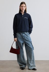 Μπλε φούτερ με φερμουάρ και γιακά, με το λογότυπο "TOMMY JEANS"· φαρδιά ανοιχτό μπλε τζιν παντελόνια cargo· κόκκινη χειροποίητη τσάντα· μαύρα παπούτσια· ουδέτερο φόντο.