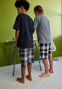 Dwoje dzieci w luźnych koszulkach: jedna granatowa, druga szara. Obydwoje w szortach w kratę—czarno-białych. Stoją boso przy zielonej ścianie.