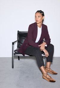 Bordeauxrotes Sakko über einem weißen T-Shirt, kombiniert mit dunklen Jeans und braunen flachen Schuhen. Auf einem schwarzen Stuhl vor einfarbigem Hintergrund sitzend.