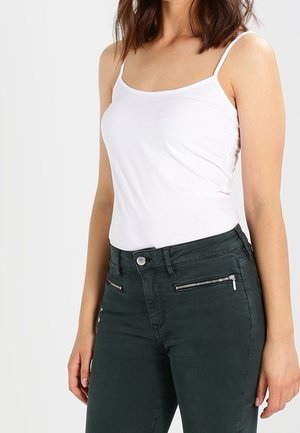 Nainen, jolla on päällään istuva valkoinen toppi ja tummanvihreät korkeavyötäröiset farkut, joissa on vetoketjutaskut edessä, seisoo yksiväristä taustaa vasten.