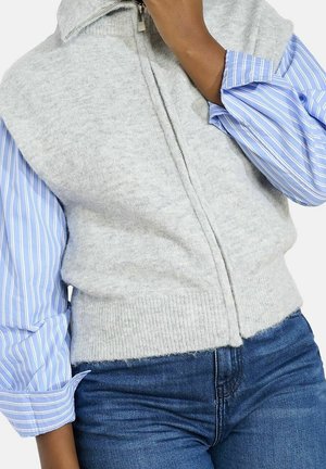 Personne portant un gilet en tricot gris clair zippé sur une chemise à rayures bleues et blanches à boutons, et un jean bleu, main près du visage.