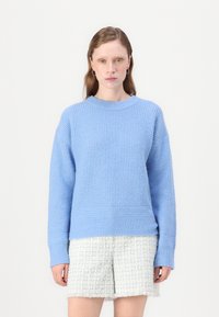 Világoskék kötött pulóver, textúrával ellátott mintával, kerek nyakkal és bordás mandzsettákkal. Off-white textúrált rövidnadrággal viselve.