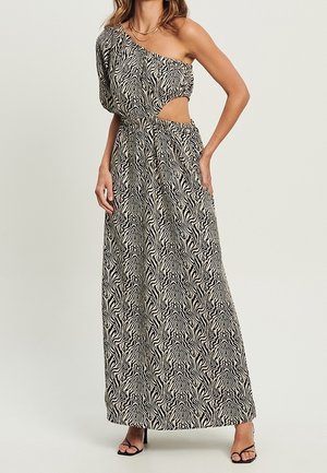 Kobieta ubrana w czarno-białą maxisukienkę na jedno ramię w zebra wzór z wycięciem w talii i czarne sandały na obcasie z paskami.