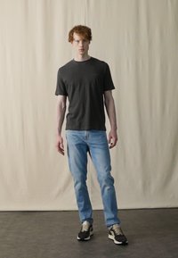 Tricou gri din bumbac, blugi deschiși la culoare și adidași negri cu accente bej. Modelul stă pe un fundal neutru.