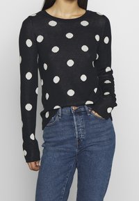 Musta pitkähihaine villapusero valkoisilla pilkuilla, pyöreä kaula-aukko ja ribbauskankaiset hihansuut, yhdistettynä korkeavyötäröisiin sinisiin farkkuihin.