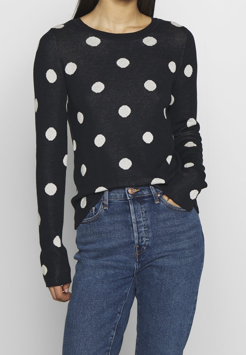 Musta pitkähihaine villapusero valkoisilla pilkuilla, pyöreä kaula-aukko ja ribbauskankaiset hihansuut, yhdistettynä korkeavyötäröisiin sinisiin farkkuihin.