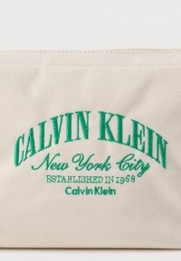 Μπεζ ύφασμα με πράσινο κεντητό κείμενο: "CALVIN KLEIN New York City ΙΔΡΥΘΗΚΕ ΤΟ 1968 Calvin Klein."