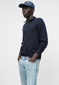 Pullover navy a trama con colletto zip, abbinato a jeans chiari strappati. Il berretto è in tessuto denim con visiera arrotondata.