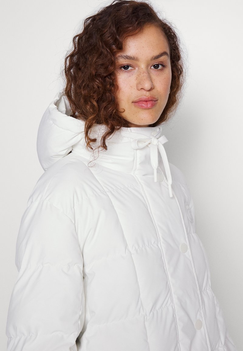 Doudoune blanche avec un col montant, une capuche à cordon et des détails à boutons-pression, présentant une texture lisse et matelassée.