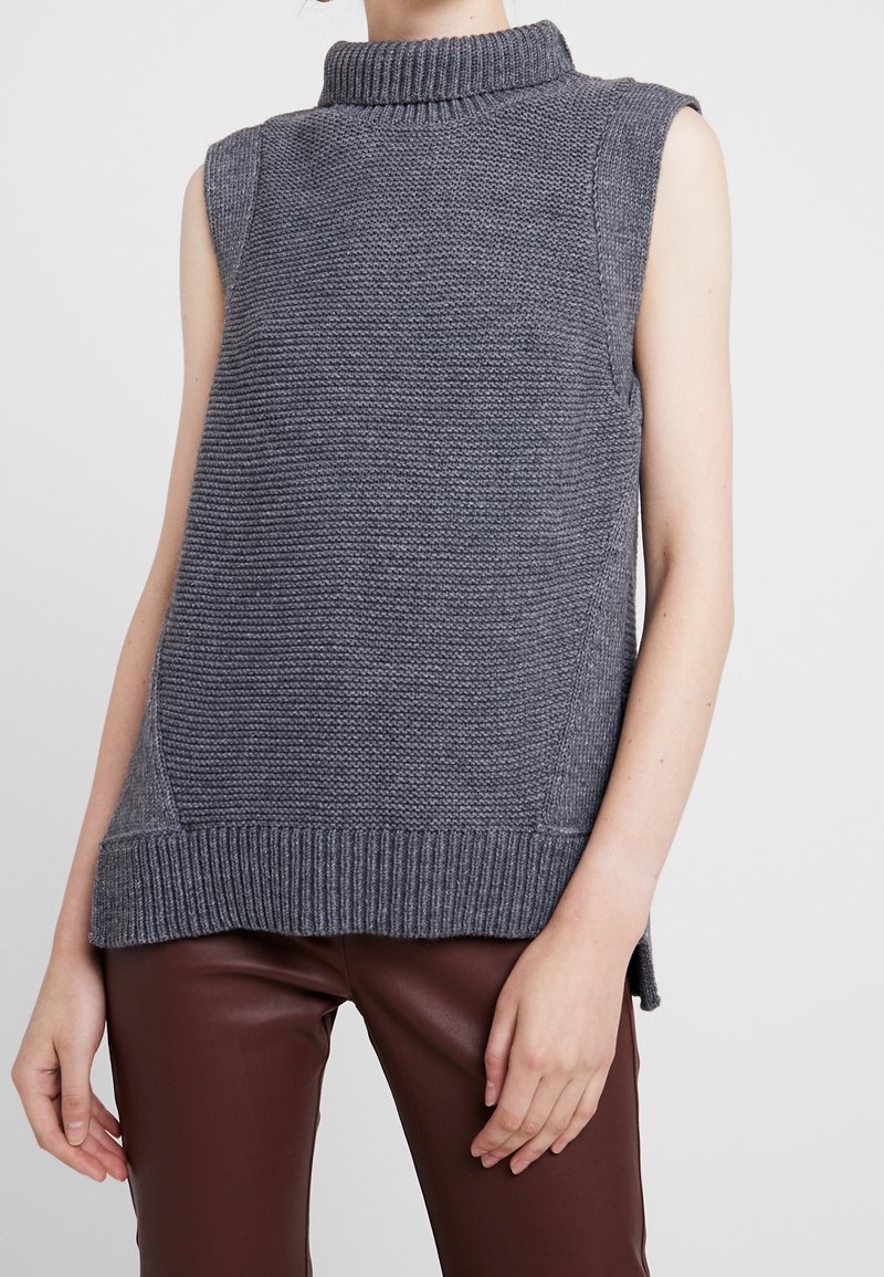 Maglione senza maniche grigio lavorato a maglia con collo alto, rifinito con orlo a costine e motivo testurizzato. Indossato sopra pantaloni di pelle marrone.