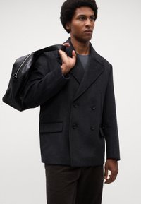 Donkerwollen dubbelzijdige peacoat met geknipte revers, voorzakken, gedragen over een grijze shirt, gecombineerd met een zwarte leren schoudertas.