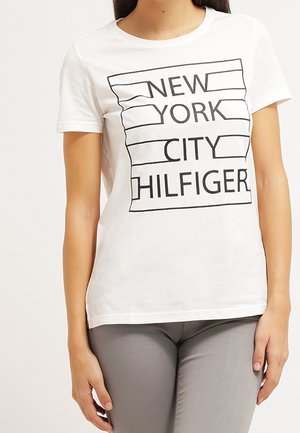 Mujer con una camiseta blanca con texto en contorno negro que dice "NEW YORK CITY HILFIGER" y pantalones grises, sobre un fondo liso.